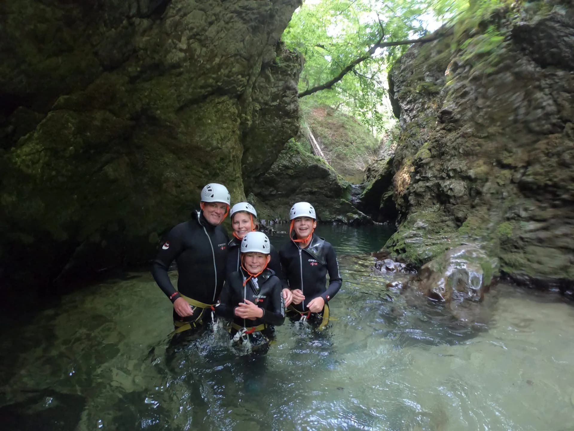 Family in wetsuits and helmets canyoning in a narrow gorge with mossy rock walls.