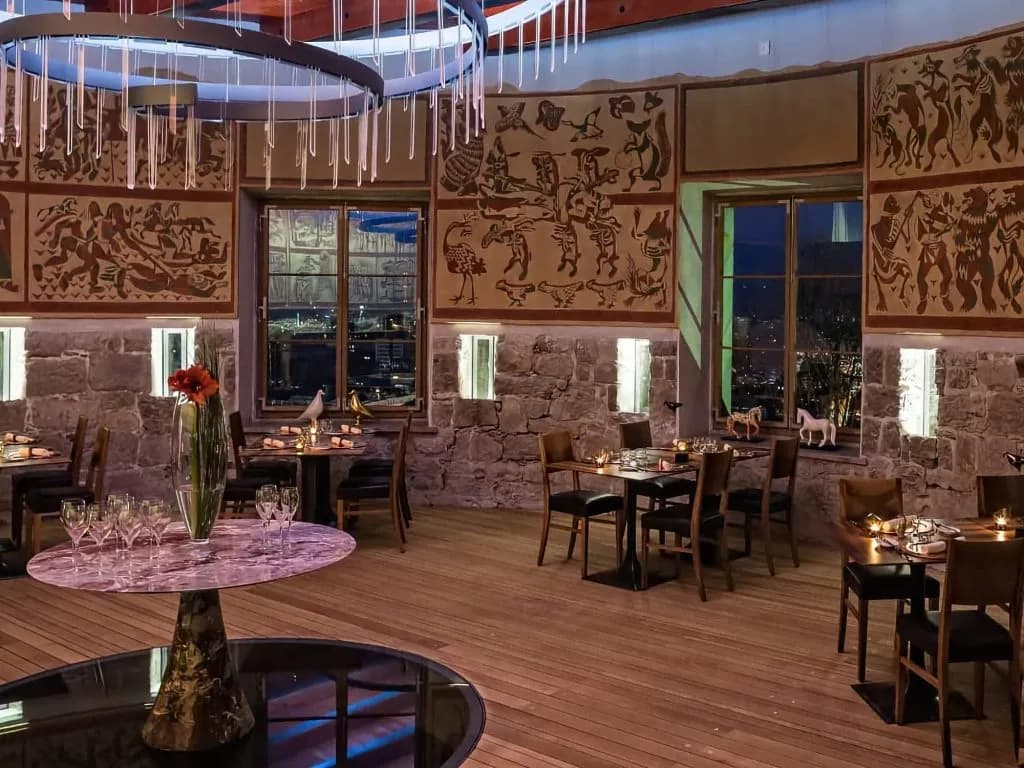 Restaurant dining room with stone walls, murals, and city lights visible through windows at night.