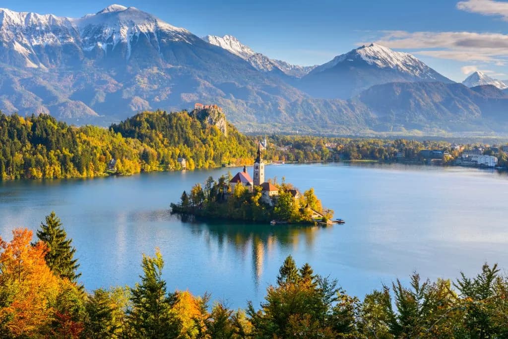 Panoramic view of Lake Bled island with church, autumn trees, and snow-capped mountains in Slovenia.