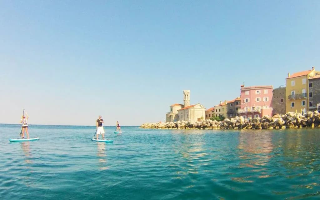 Stand-up paddleboarding near colorful coastal buildings and a stone church in Piran, Slovenia.